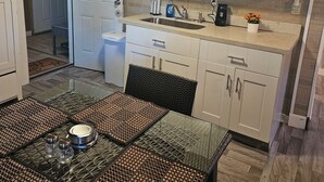 Cocina pequeña con armarios blancos, un lavamanos y una zona de comedor con mesa de vidrio y individuales tejidos.