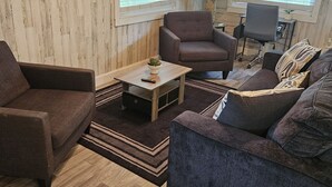 Una sala de estar acogedora con un televisor de pantalla plana, dos ventanas con persianas, un sillón gris, un sofá, una mesa de centro con una planta y un escritorio con una silla.