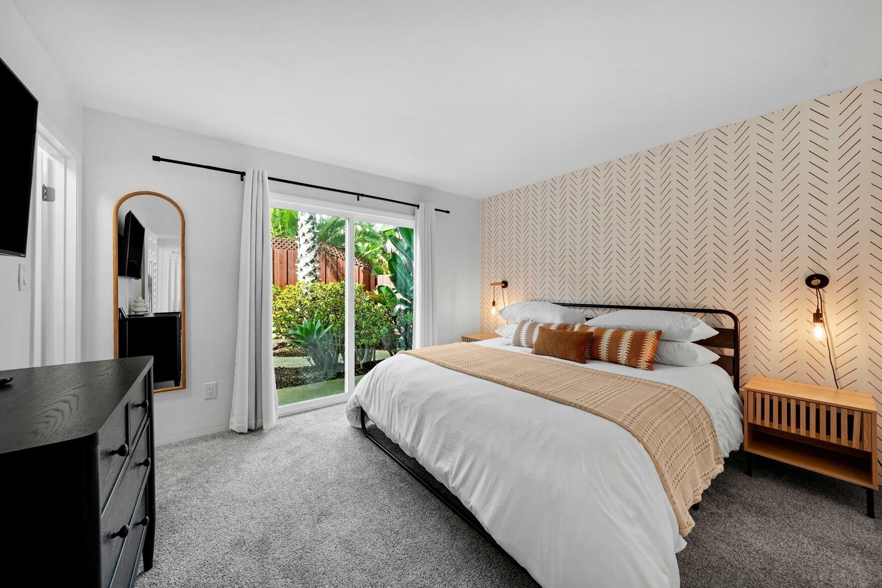 A bedroom with a bed, a dresser, a TV, and a sliding glass door leading to a garden.