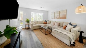 A modern living room with a sectional sofa, a coffee table, a TV, and a large window with blinds. - Luxe & Spacious Retreat - Steps to Beach Game Room (Encinitas)