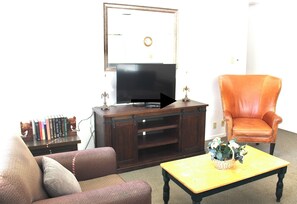 Ein Wohnzimmer mit Fernseher, einer Couch, einem Sessel, einem Couchtisch und einem Bücherregal.