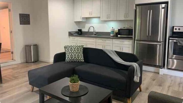 Een moderne woonkamer met een zwarte bank, een klein bijzettafeltje en een keuken met witte kasten en roestvrijstalen apparatuur.