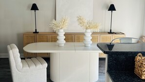 Un espace repas moderne avec une table blanche, deux vases blancs contenant des fleurs séchées, une chaise blanche à revêtement et une lampe noire.