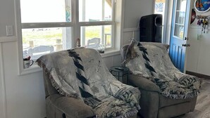 A room with two grey armchairs covered with patterned blankets, a blue door, and a window with a view of the outdoors.