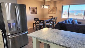 A kitchen with a stainless steel refrigerator, a dining table with chairs, a sofa, and a large window with a view of the water. - Escape to Spicer and enjoy stunning lake views from this comfortable 2-bedroom! (Spicer)