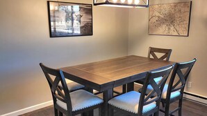 A dining area with a wooden table and four chairs, a hanging light fixture, and two framed pictures on the wall. - Escape to Spicer and enjoy stunning lake views from this comfortable 2-bedroom! (Spicer)