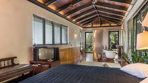 Une chambre à coucher avec un grand lit, un banc en bois, une télévision et une salle de bain visible à travers une porte ouverte.