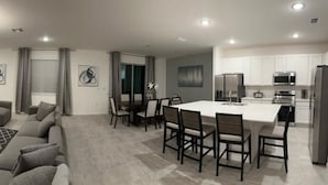 A modern living room with a flat-screen TV, grey sofas, a dining area with a white table and chairs, and a kitchen with white cabinets and appliances. - Grand Resort Home in Resort with Pool (Kissimmee)