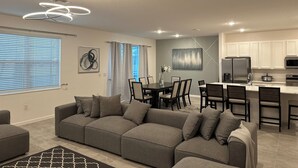 A modern living room with a gray sectional sofa, a dining area with a table and chairs, a kitchen with white cabinets and stainless steel appliances, and a ceiling with recessed lighting. - Grand Resort Home in Resort with Pool (Kissimmee)