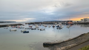 A harbor with numerous boats docked, a stone wall, and a distant view of a building with a sunset in the background.
