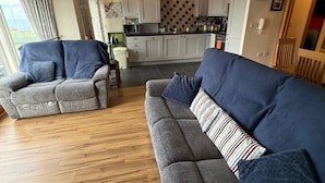 A modern living room with a grey sofa, a smaller grey armchair, and a wooden floor. A kitchen with white cabinets and a tiled backsplash is visible in the background.