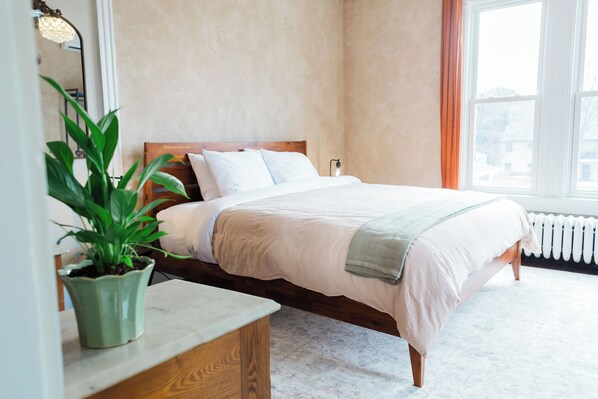 A neatly made bed with a wooden headboard, a potted plant on a bedside table, and a window with curtains.