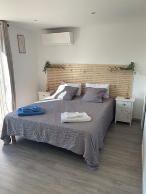 Een net opgemaakt bed met een grijze spreidek en kussens, een nachtkastje met een lamp, en een wandairconditioner.