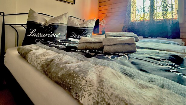 A neatly made bed with a patterned bedspread and two pillows with 'Luxurious' text, a framed floral artwork on the wall, and a window with decorative curtains. - House Bernstein, Apartman 3 (Haus im Ennstal)