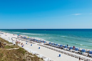 A beach with rows of striped beach umbrellas and lounge chairs, a pier in the background, and a clear blue sky.