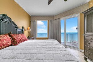 A hotel room with a large bed, a dresser, a nightstand with a lamp, and a balcony with a view of the sea. - Grandview East 1604 (Panama City Beach)