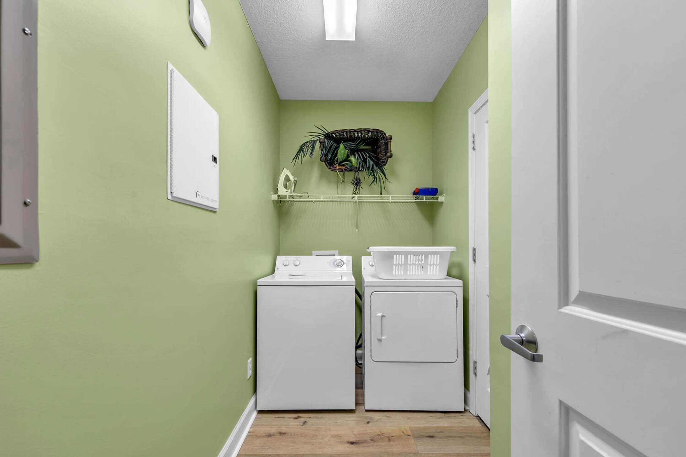 A laundry room with two white washers and dryers, a shelf with a plant, and a door on the right.