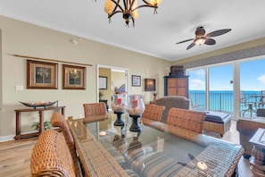 A spacious living room with a glass-top dining table, wicker furniture, and a view of the ocean through large windows. - Grandview East 1604 (Panama City Beach)