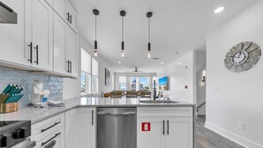 A modern kitchen with white cabinetry, a stainless steel dishwasher, pendant lighting, and a large clock on the wall. - Golden Seashell East (Panama City Beach)