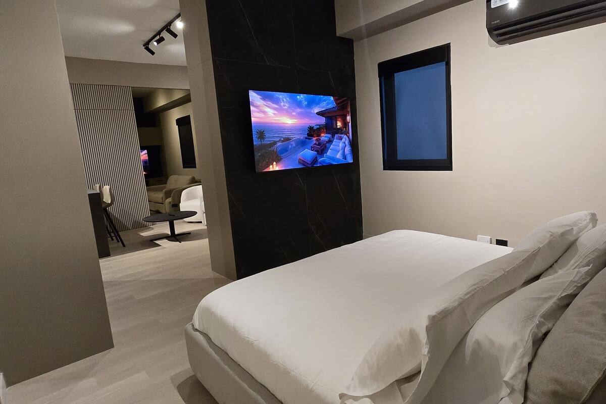 A modern hotel room with a large bed, a TV mounted on the wall, and a view of the beach through the window.