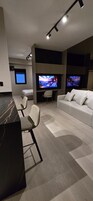 Una sala de estar moderna con un sofá, dos sillones, una mesa de centro y un mueble pared para televisión con dos pantallas.