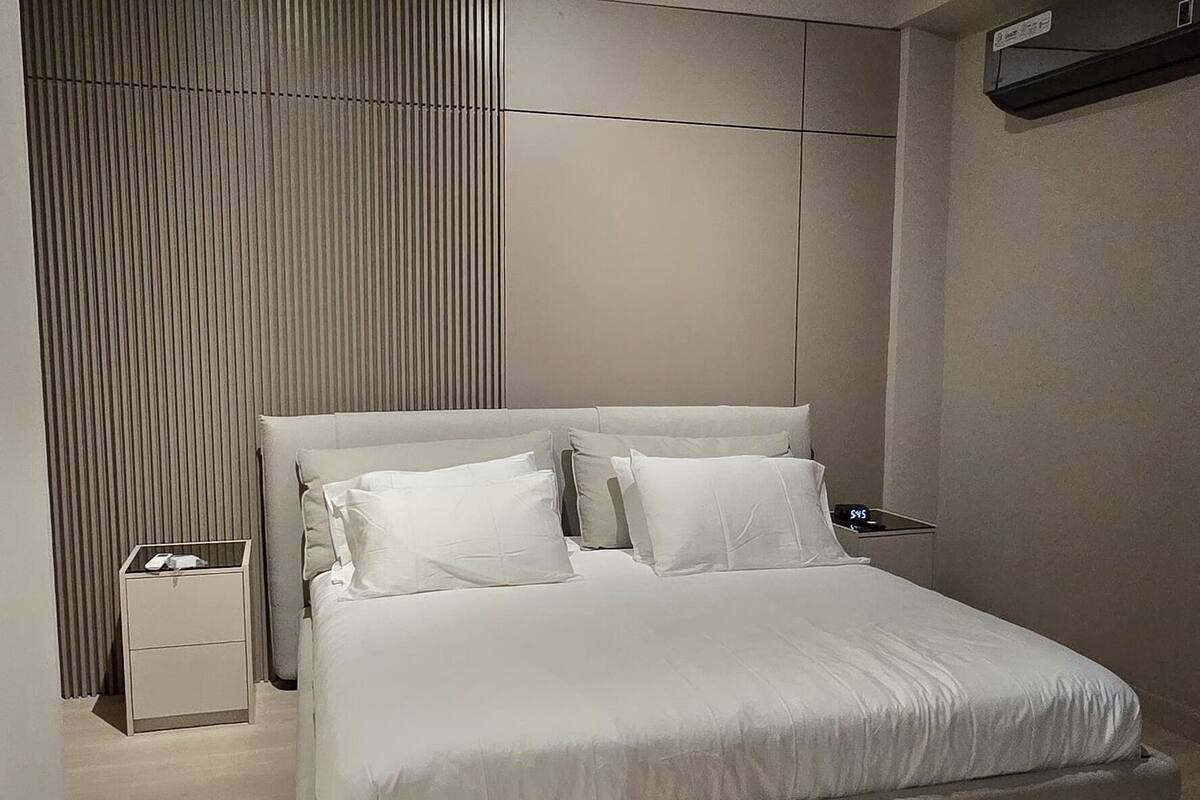 A modern bedroom with a large bed, a bedside table, a wall-mounted air conditioner, and a ceiling light fixture.