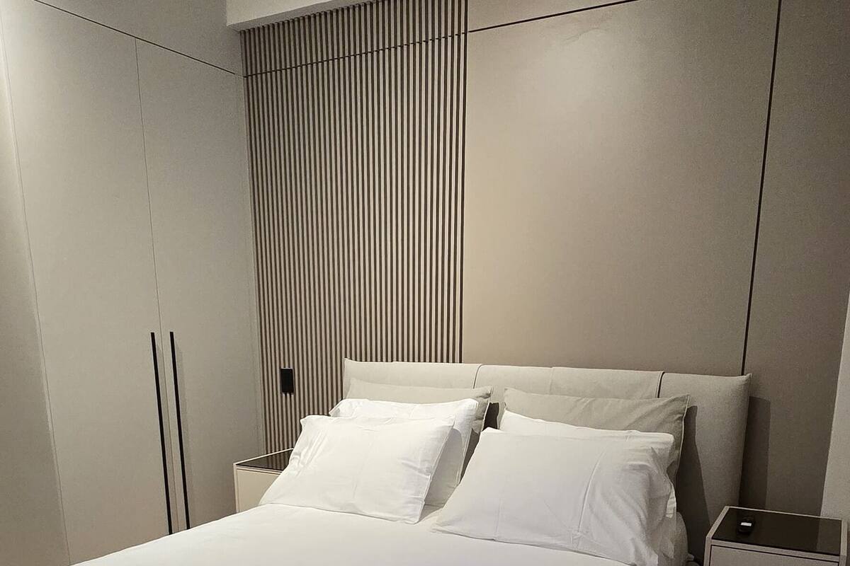 A modern bedroom with a large bed, a nightstand, a wardrobe, and a wall-mounted panel.