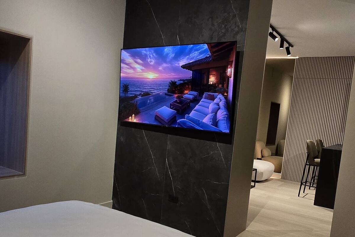 A modern bedroom with a large bed, a flat-screen TV mounted on a dark wall, and a small desk area with a chair.