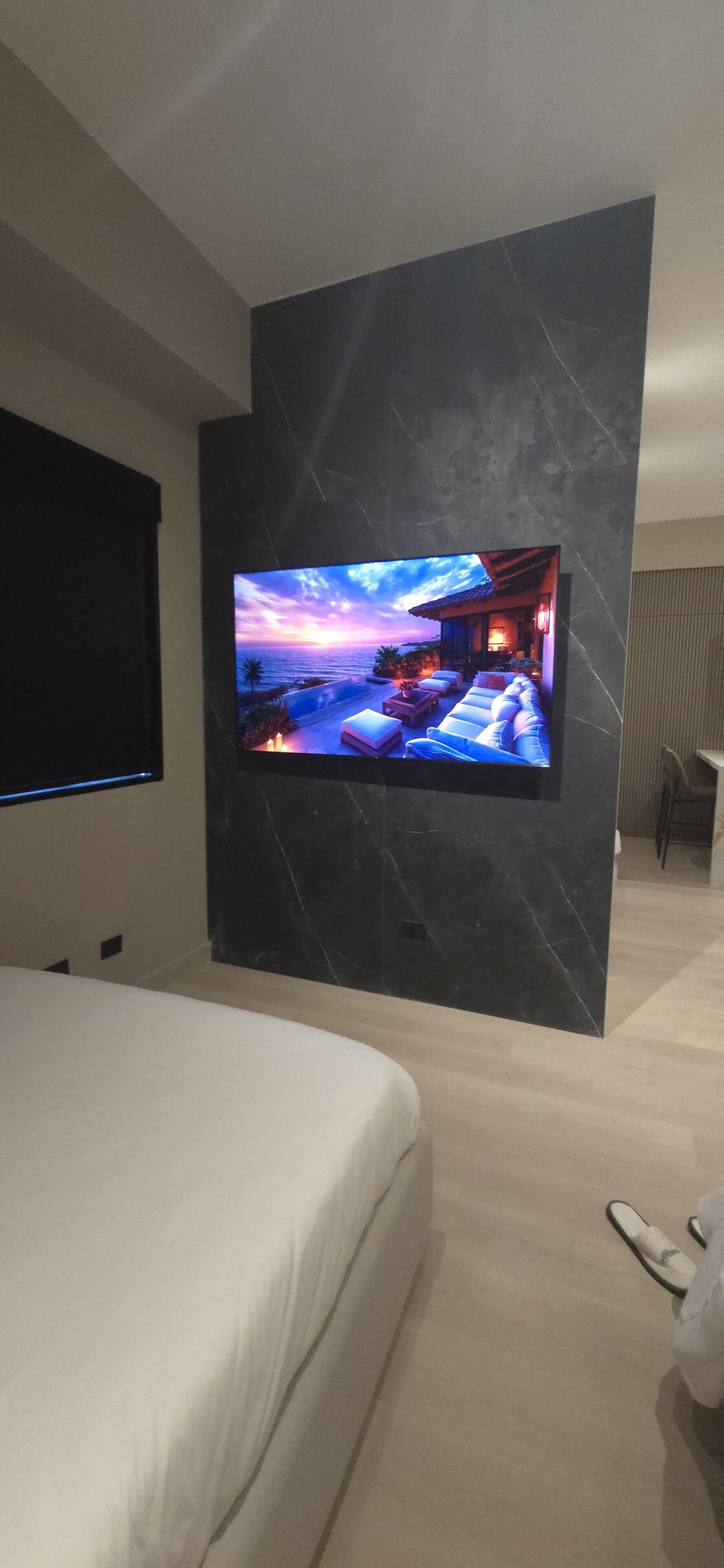 A modern bedroom with a large flat-screen TV mounted on a marble wall, a bed with white bedding, and a pair of slippers on the floor.