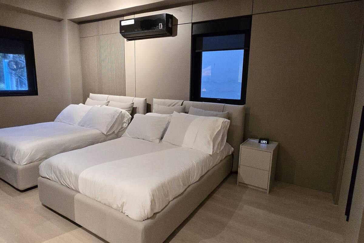 A modern hotel room with two beds, a small bedside table, a TV mounted on the wall, and a window providing a view.