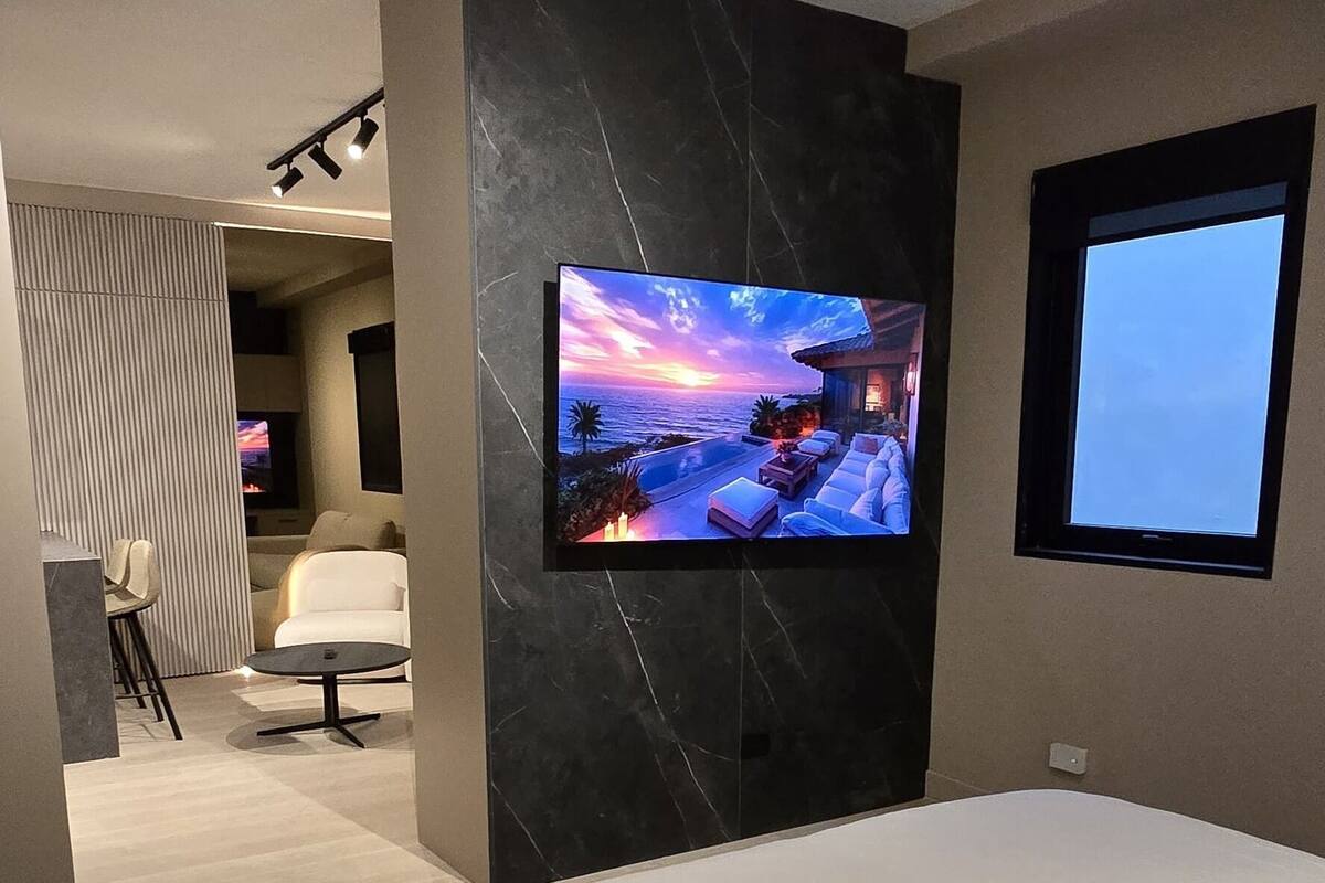 A modern hotel room with a large bed, a flat-screen TV mounted on a marble wall, and a view of the sea through a window.