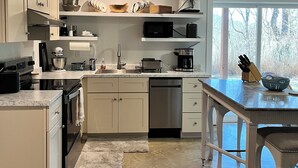 Cozinha moderna com armários brancos, lava-louças, fogão e uma janela grande com vista para árvores.