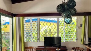 Un comedor con mesa y sillas de madera, televisor y vista al cielo azul y vegetación a través del ventanal.
