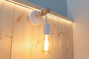 A wall-mounted light fixture with a clear glass bulb, attached to a wooden panel with a white circular base. - Velis - art & hospitality (Posada)