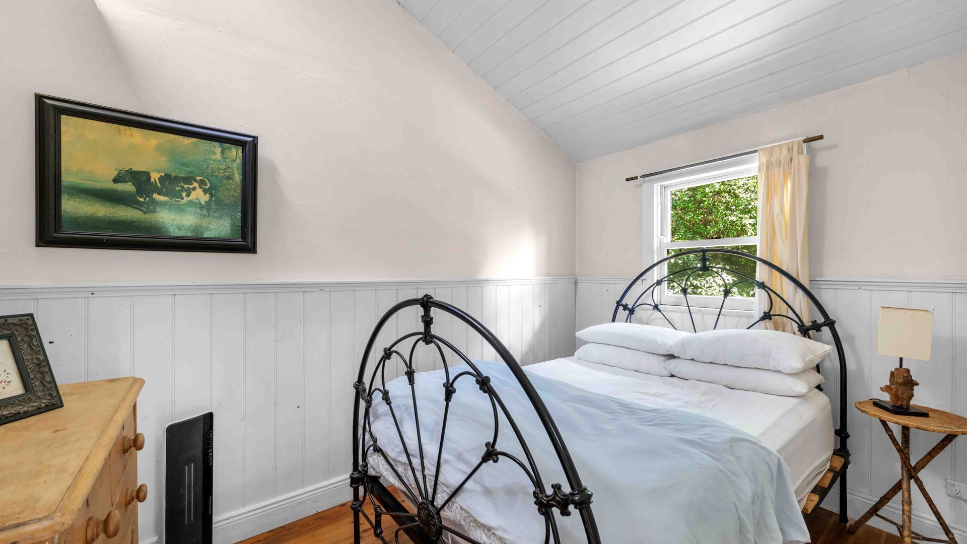 A bedroom with a curved ceiling, a bed with white linens, a painting of a cow, a wooden dresser, and a window with a view of greenery.