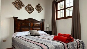 Un dormitorio con cama de madera, colcha acolchada, decoración mural, una ventana con cortinas y una mesita de noche con lámpara.