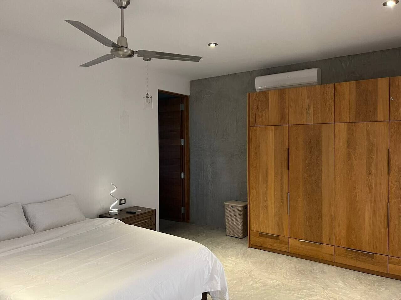 Une chambre à coucher comprenant un lit, une table de chevet, un ventilateur de plafond, une armoire et une porte donnant sur une autre pièce.