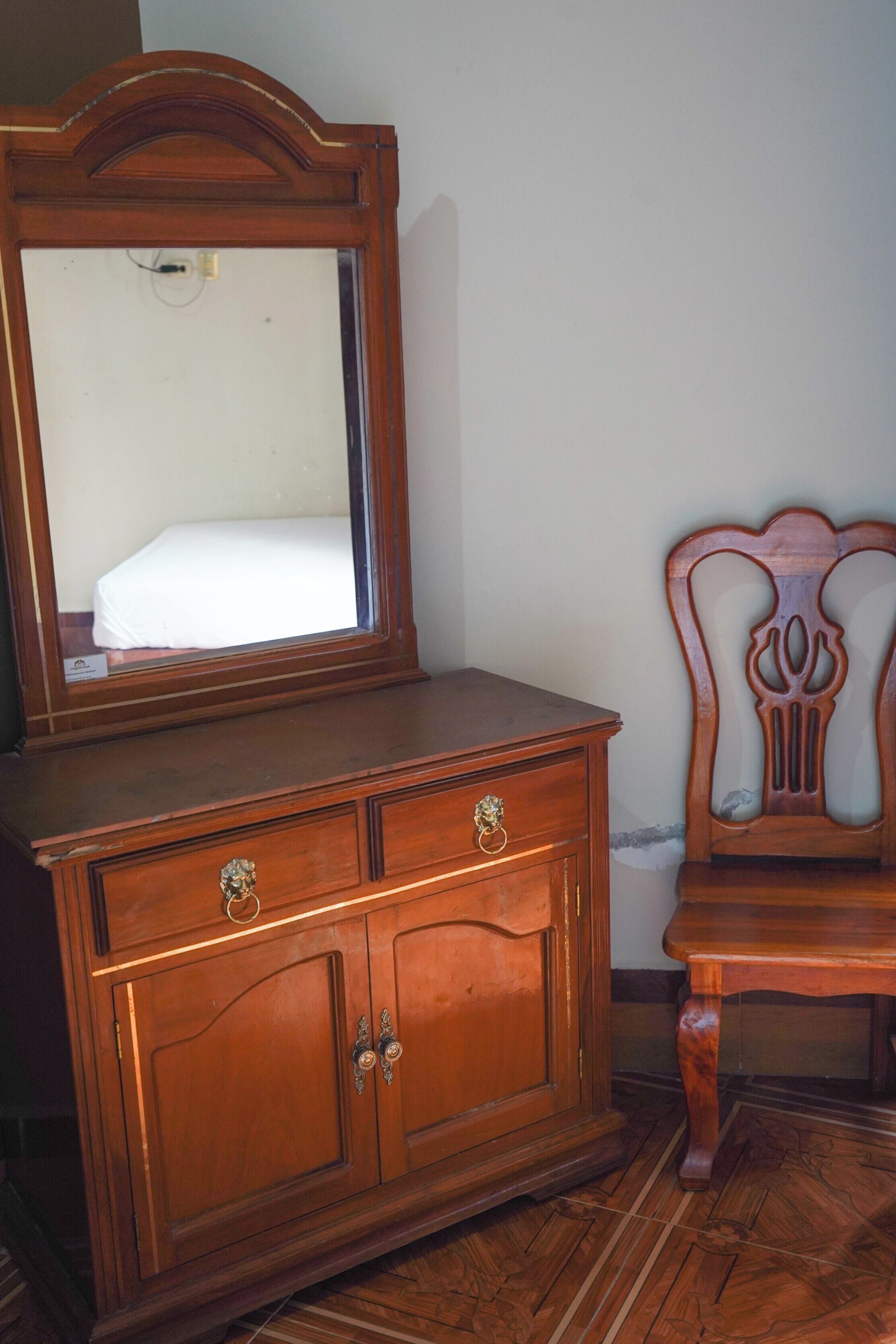 A wooden dresser with a mirror and drawers, a chair with a carved backrest, and a patterned wooden floor.