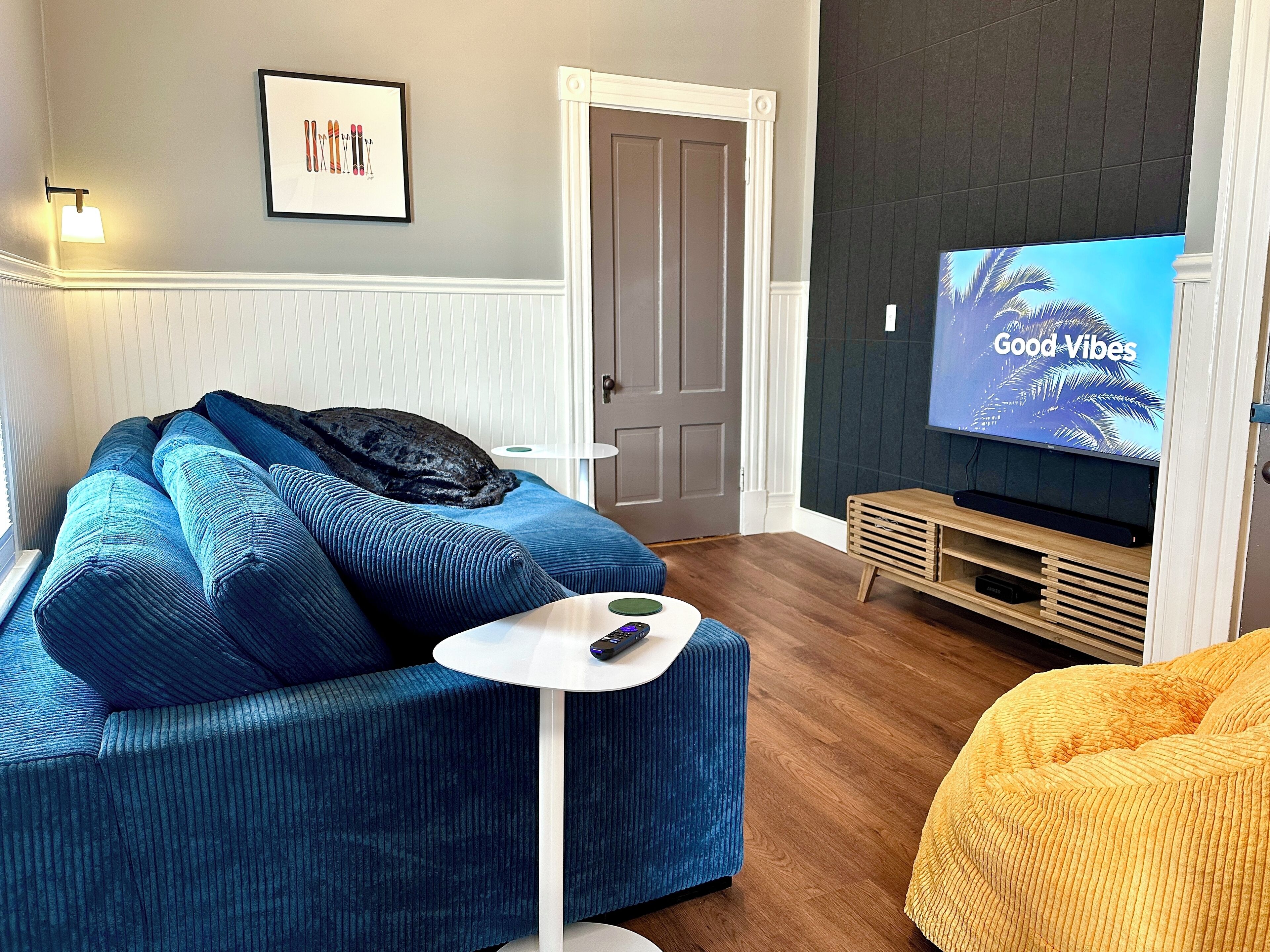 Un salon avec un canapé bleu, une petite table blanche, un téléviseur diffusant l’émission « Good Vibes », et une porte visible au fond.