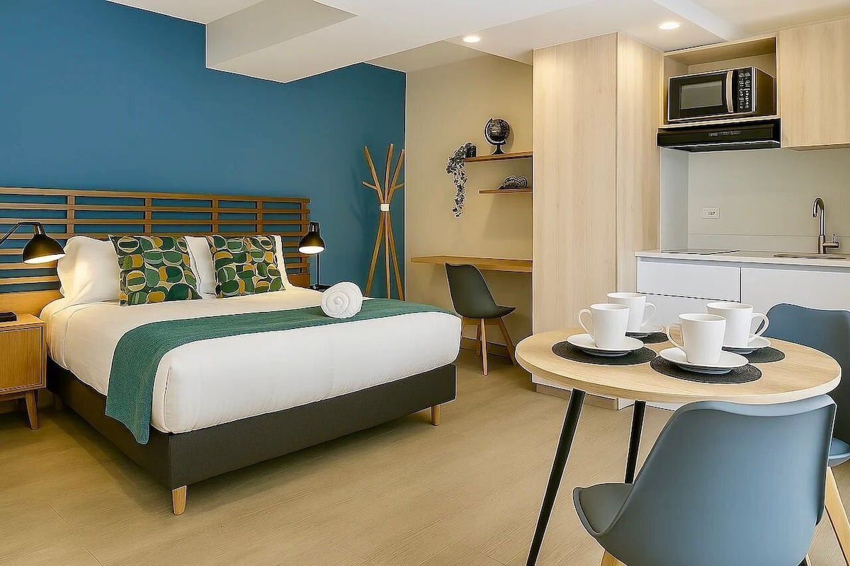 A hotel room with a bed, bedside tables, a desk, a chair, a dining table with cups, and a microwave in the kitchen area.