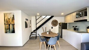Cocina moderna con horno empotrado, microondas y refrigerador. Hay un comedor con una mesa redonda y cuatro sillas. Una escalera con barandal negro conduce a otro nivel.