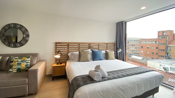 Habitación de hotel con una cama grande, un cabecero de madera, un espejo redondo, una mesita de noche, una lámpara, un sofá y vistas de edificios a través de un ventanal amplio.