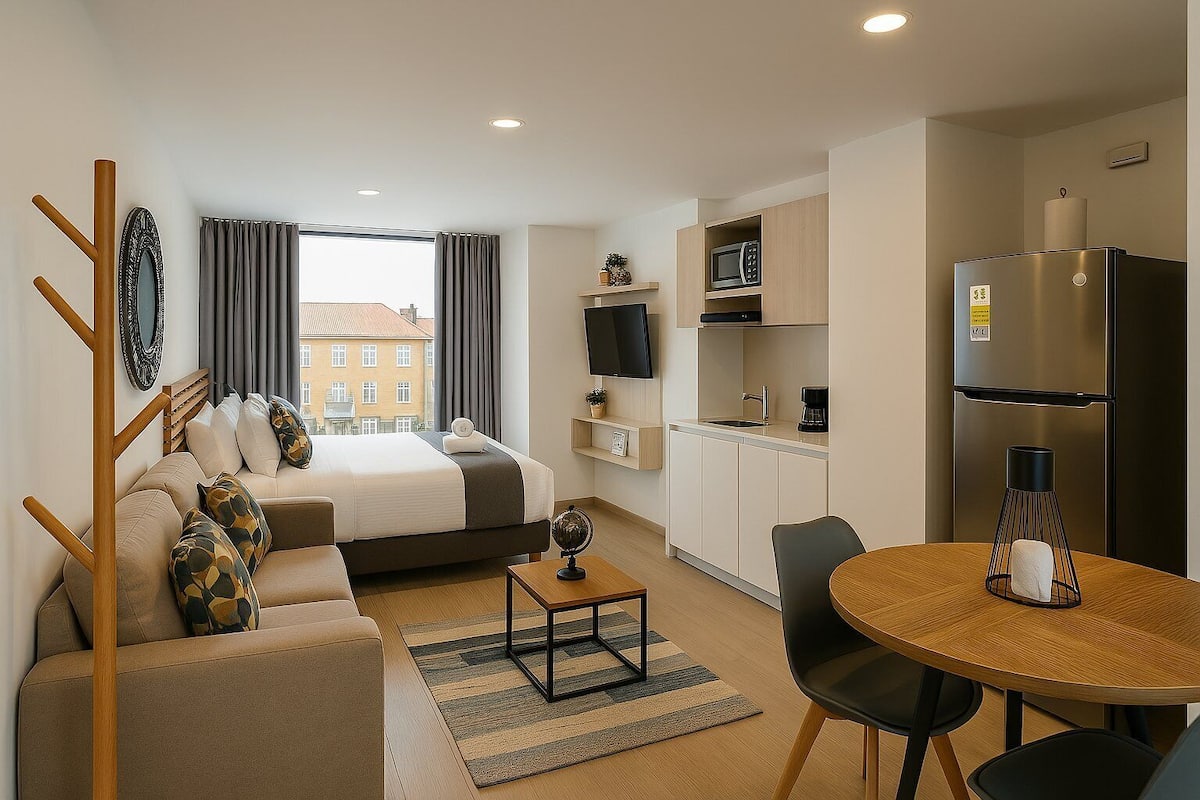 A compact hotel room with a bed, a sofa, a dining table, a kitchen area with a microwave and refrigerator, and a TV.