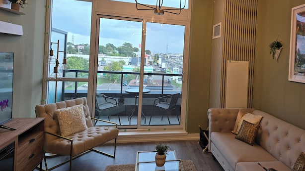 A modern living room with a sofa, armchair, and a dining table. There is a large window with a view of the city. - Modern Whitby 3 Bedroom Condo, close to the 401, Trafalgar Castle and Toronto! (Whitby)