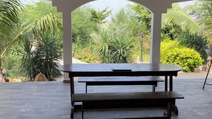 A covered outdoor area with a wooden bench and a table, surrounded by lush greenery and a view of the outdoors.