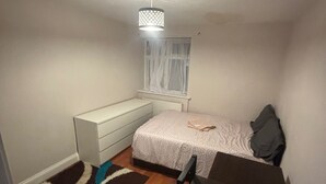 A small, dimly lit room with a single bed, a desk, and a chair. There is a window with sheer curtains, a radiator below the window, and a patterned rug on the floor. - Stylish 1-bed Apartment- Sleeps 3 w/ Central Links (Pinner)