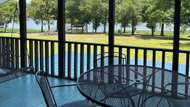 Un espacio al aire libre techado con mesa y sillas, ventilador de techo y vista a un lago rodeado de árboles.