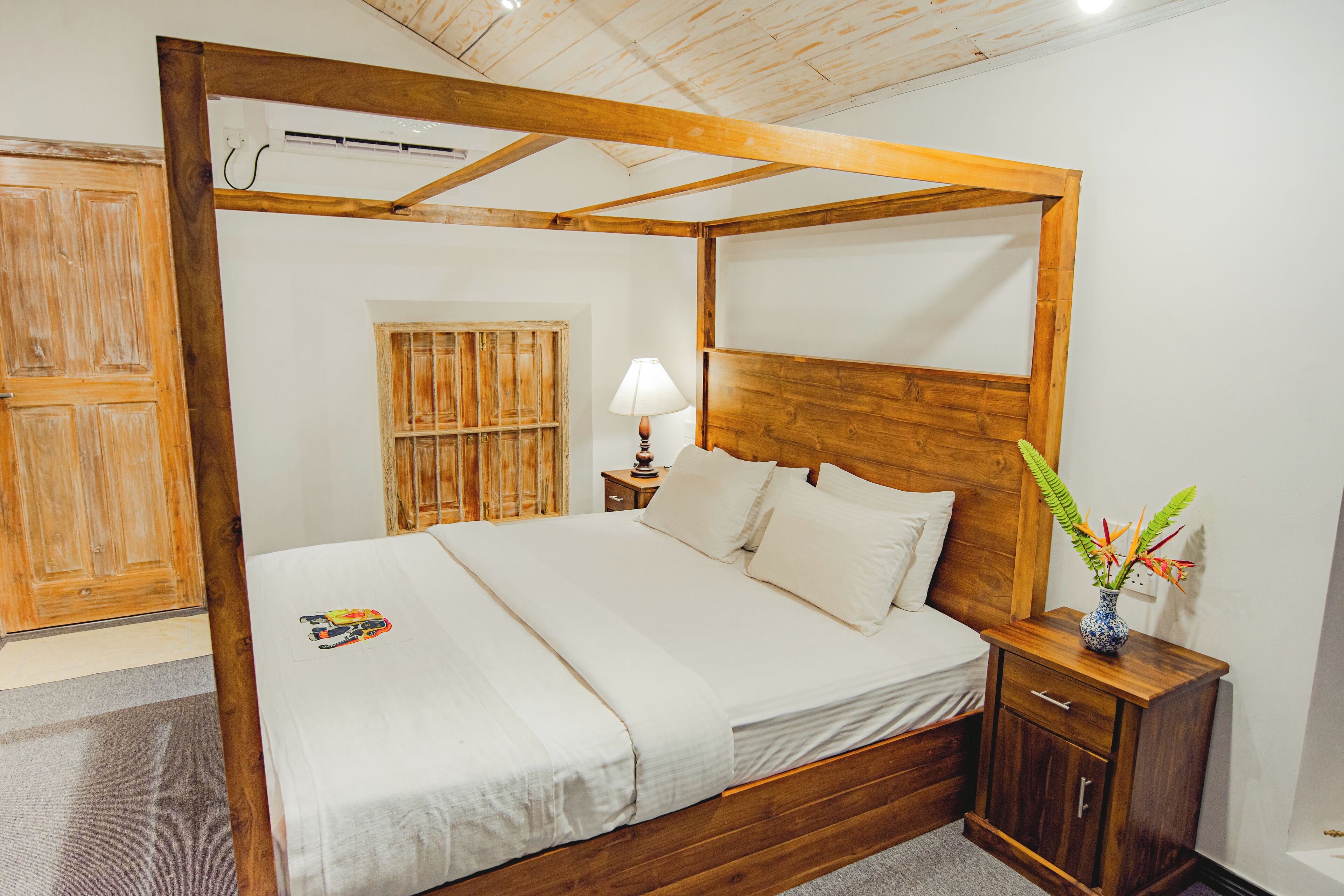 Una cama con dosel de madera, con colchón y almohadas blancos, una mesita de noche con lámpara y un jarrón pequeño con flores.