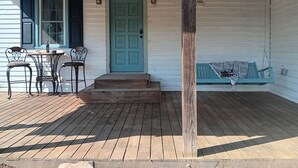 A wooden porch with a green door, two chairs, a table with a bottle, and a swing.
