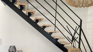 Uma escada moderna com guarda-corrimão preto e degraus de madeira, uma cadeira de vime trançado, um console de madeira com gavetas e um candeeiro suspenso.